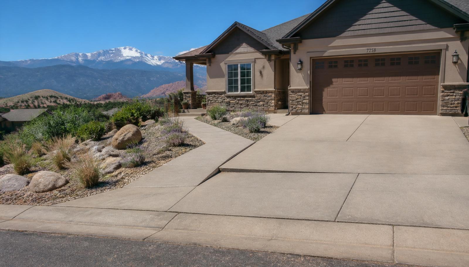 A Colorado Springs residential home with a concrete driveway, front sidewalk, and side patio showing subtle signs of concrete settlement, xeriscape landscaping and Pikes Peak with the Front Range foothills in the background